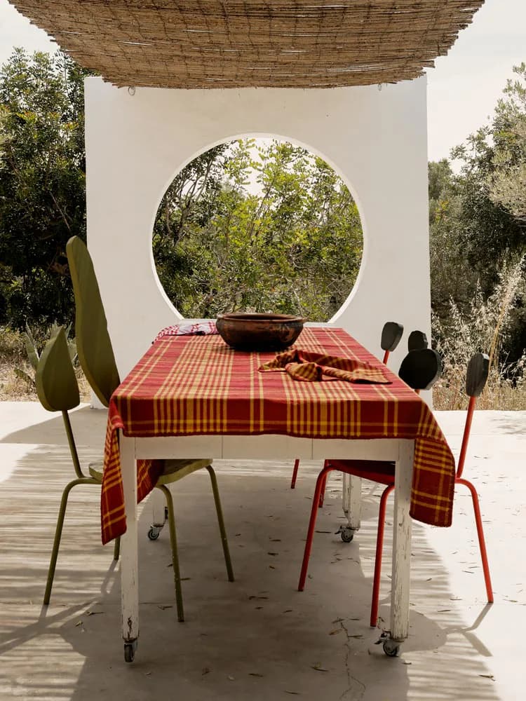 Minimalist outdoor dining installation featuring a white wall with circular aperture framing vegetation, red checkered tablecloth, and sculptural seating by Viso Project.