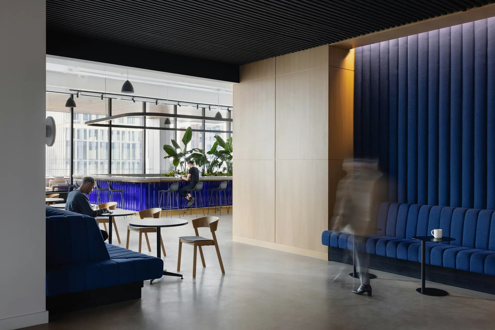 Modern office lounge with blue velvet seating, cream-colored walls, dark ceiling, and blue vertical blinds; minimalist interior design by Vocon.