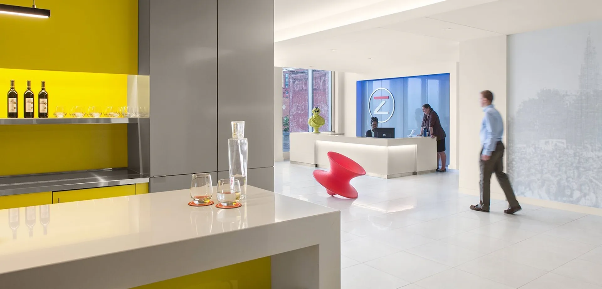 Modern office reception area with yellow and gray kitchen, white floors, and people in the background by Vocon.