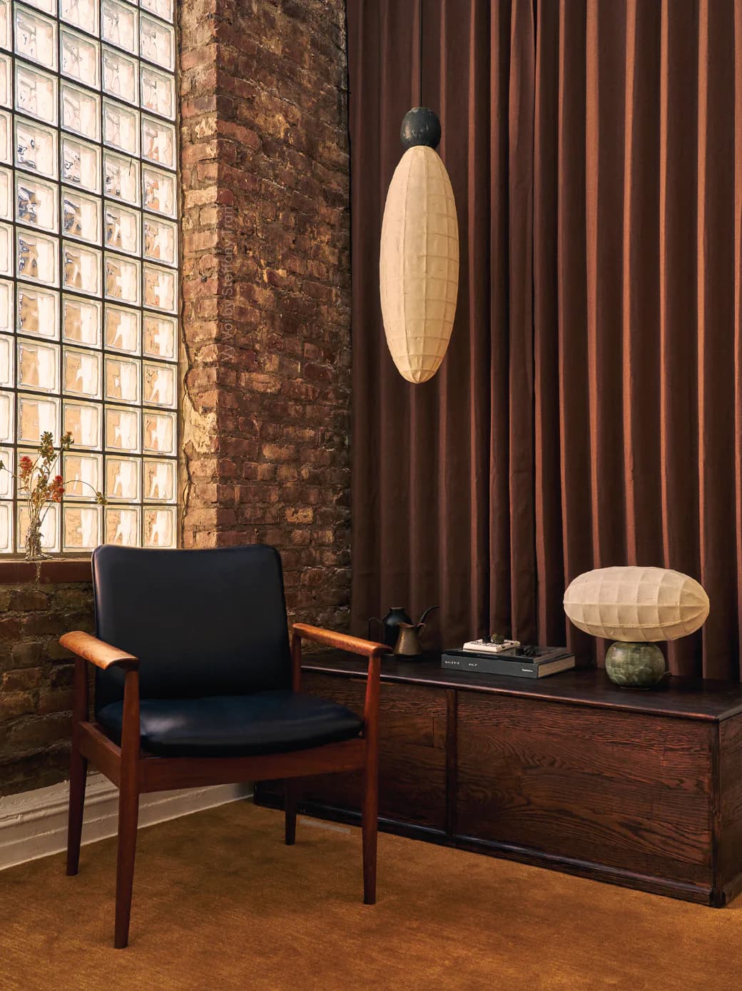 Mid-century modern lounge with brick wall, wood-framed chair, pendant lighting, and warm earthy tones by Vy Voi.