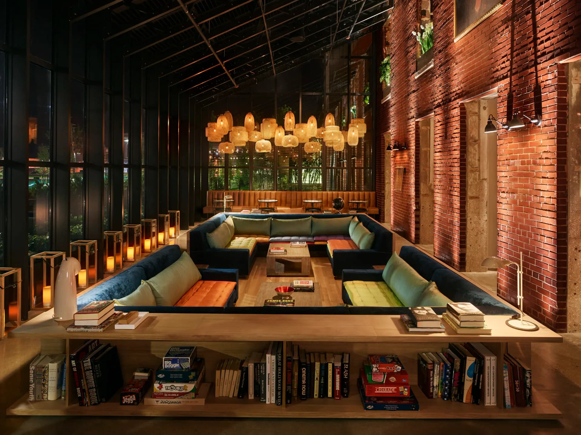 Narrow lounge with black seating, orange accents, warm pendant lighting, exposed brick walls, and built-in bookshelves by WALD Studio.