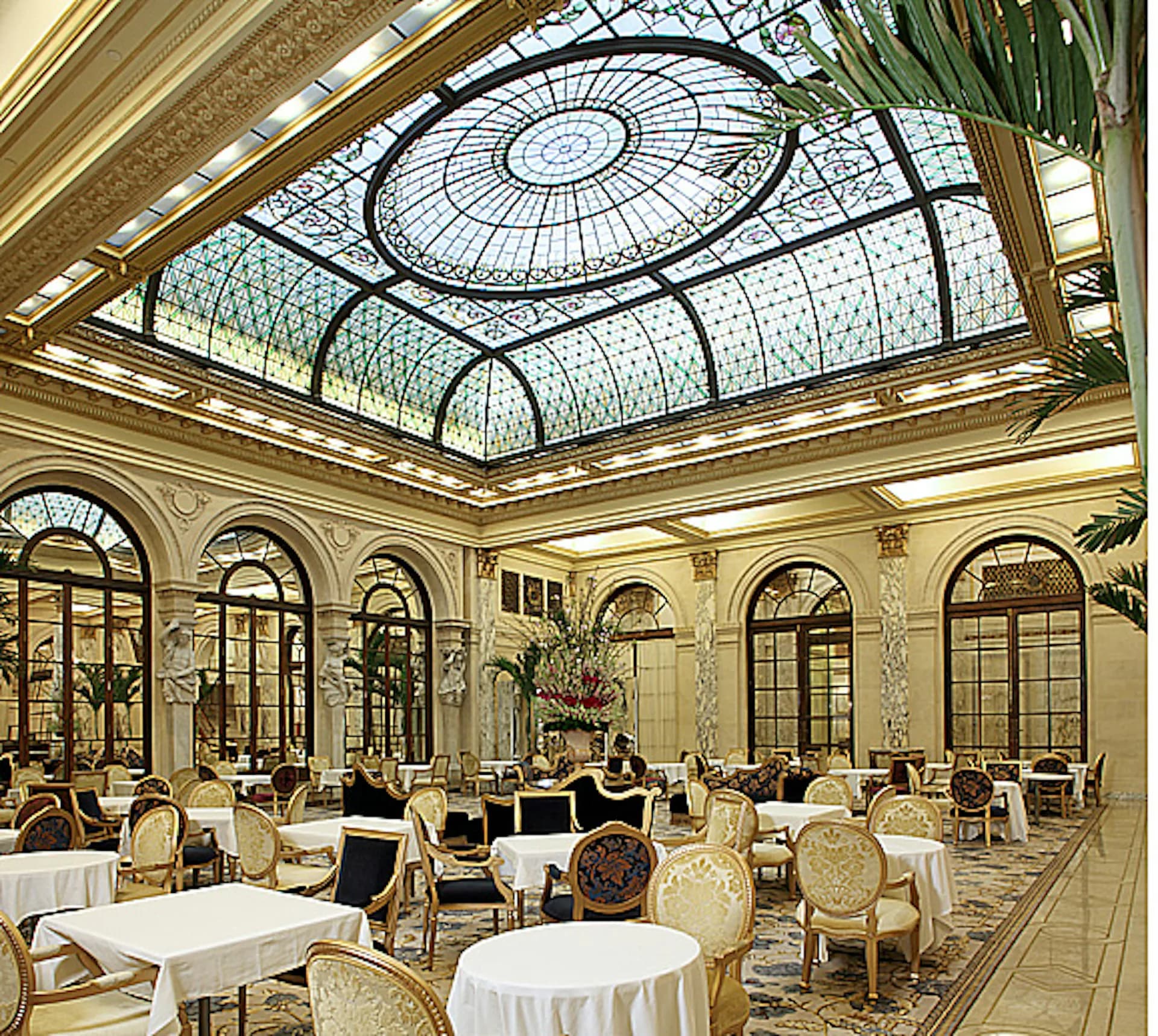 Grand ballroom with ornate cream-colored walls, arched windows, and elaborate glass-paneled skylight ceiling with geometric patterns, set with round dining tables by Walter B. Melvin Architects.