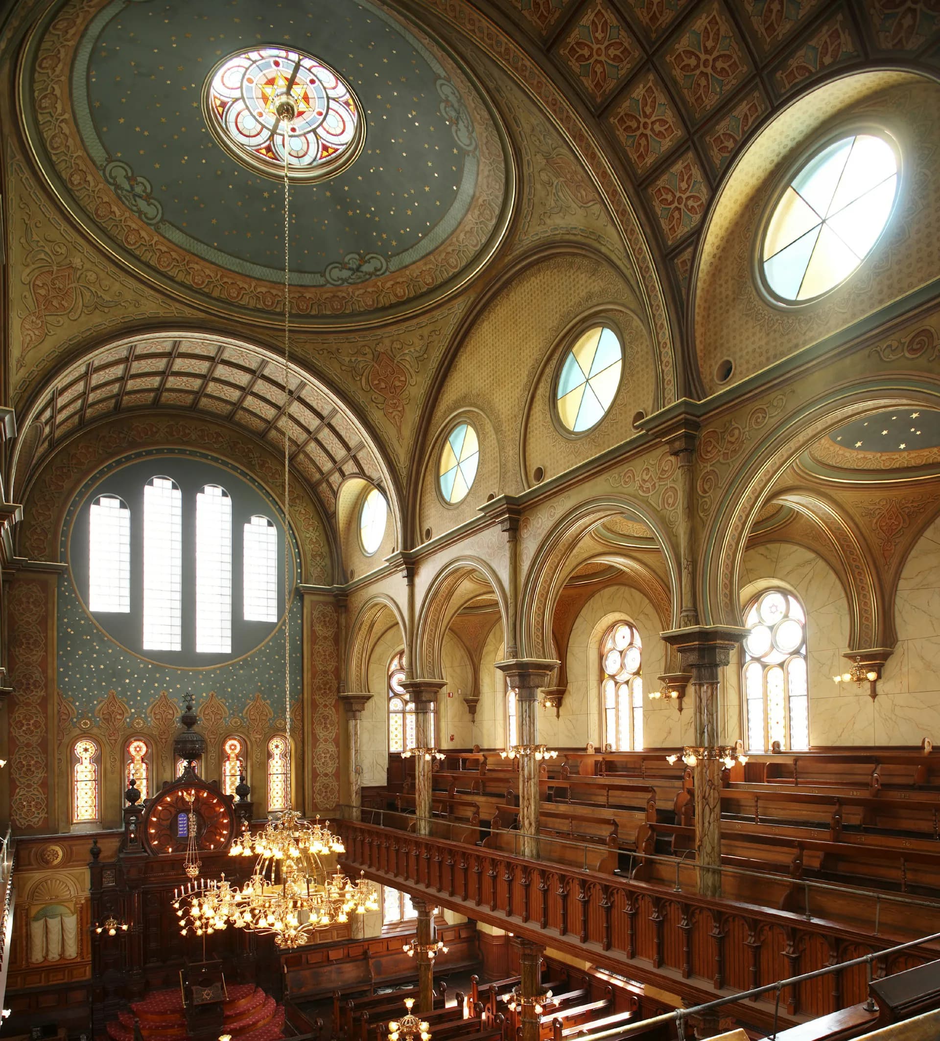Ornate church interior with barrel-vaulted ceiling, decorative tile work, wooden pews, arched windows with natural light, and hanging chandelier by Walter Sedovic Architects.