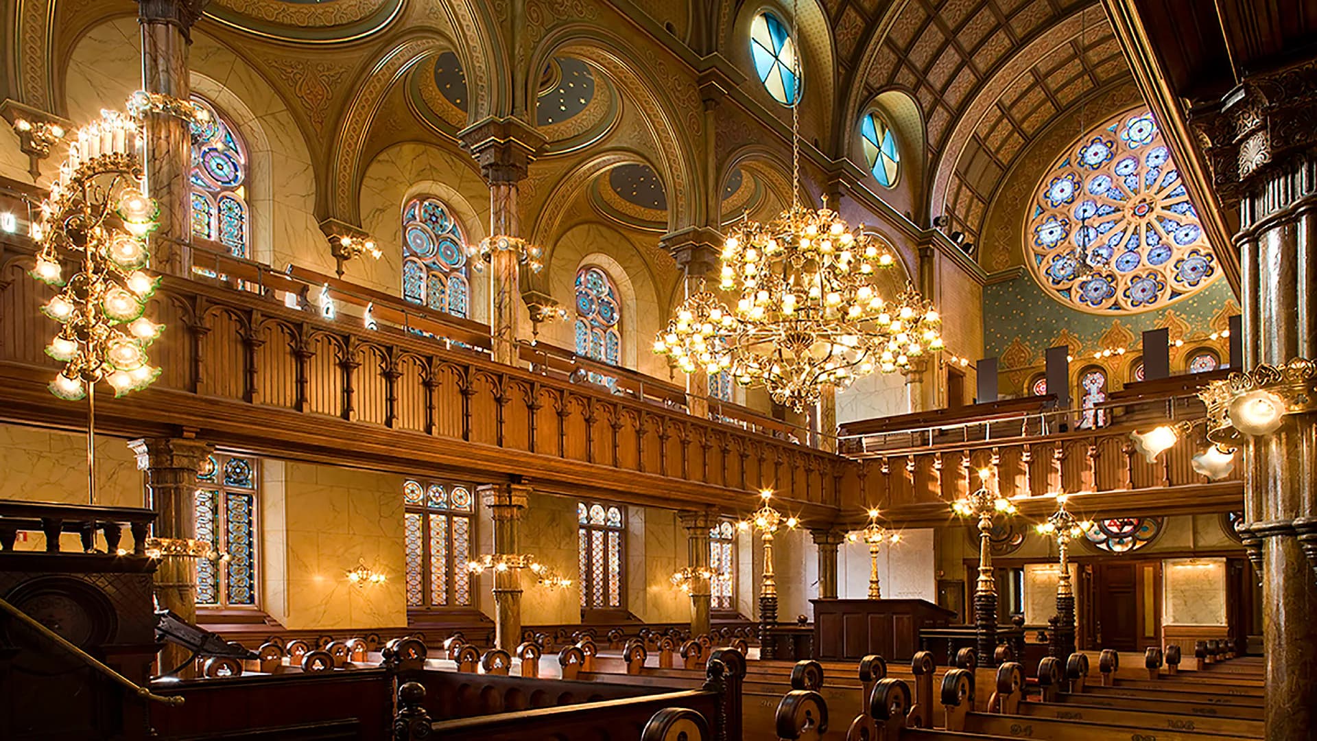 Grand synagogue interior with ornate coffered ceiling, stained glass windows, crystal chandeliers, and rich wood detailing by Walter Sedovic Architects.