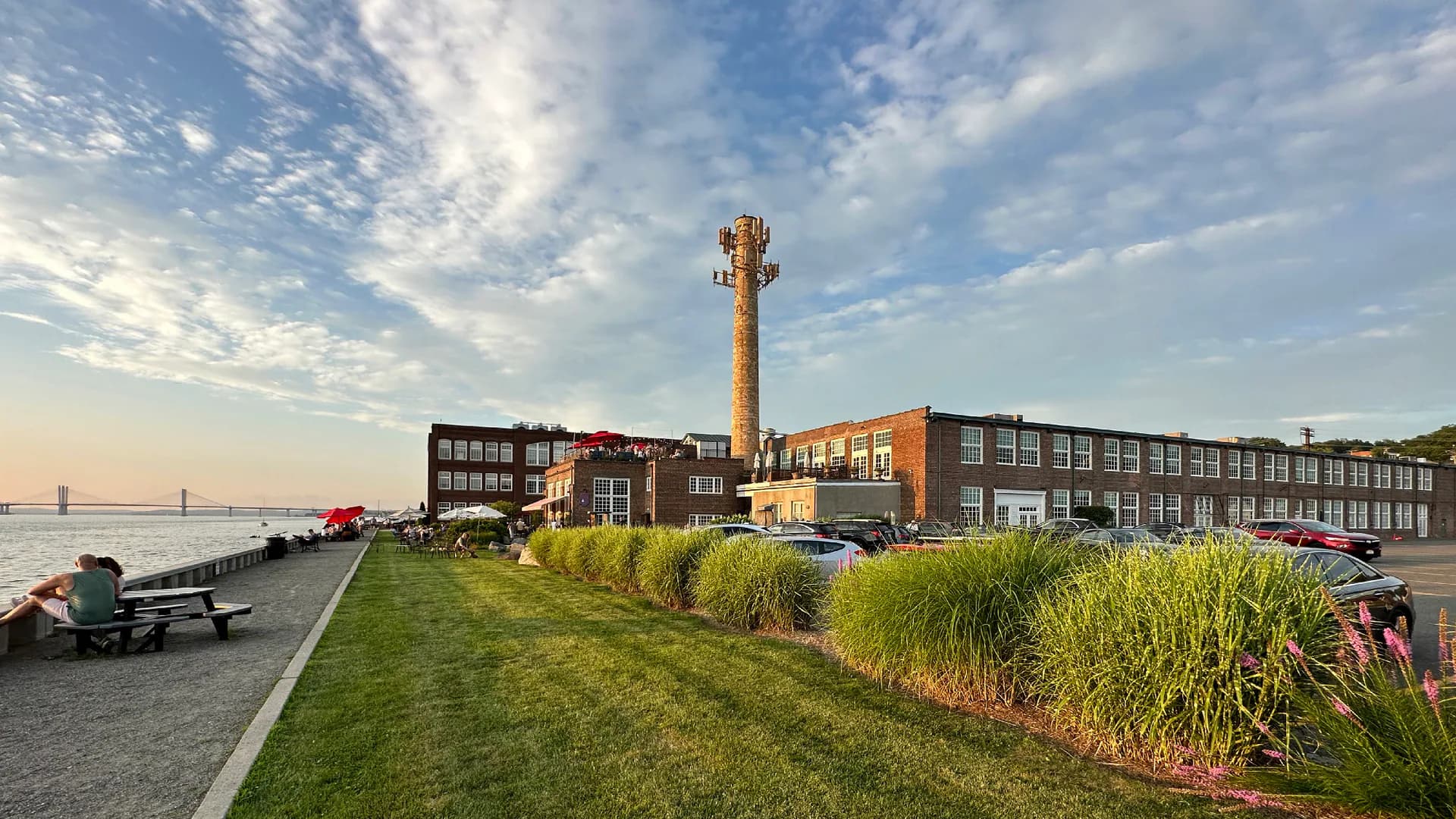 Waterfront industrial complex with brick tower, modern pavilion buildings, and native plantings along landscaped green slope by Walter Sedovic Architects.