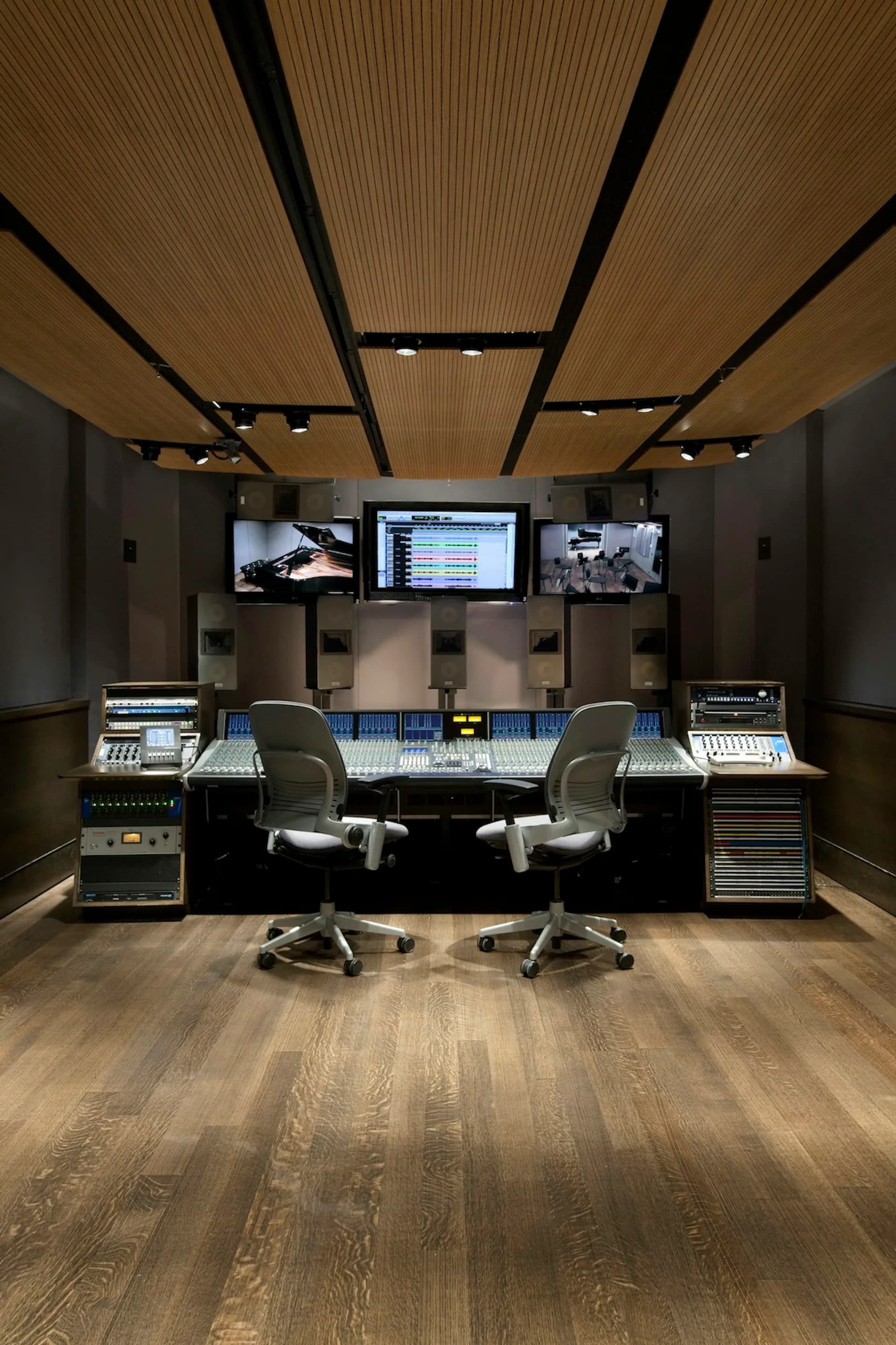 Professional recording studio control room with curved wooden ceiling panels, dual workstations with mixing consoles, three display monitors, and warm wood flooring by Walters-Storyk Design Group.