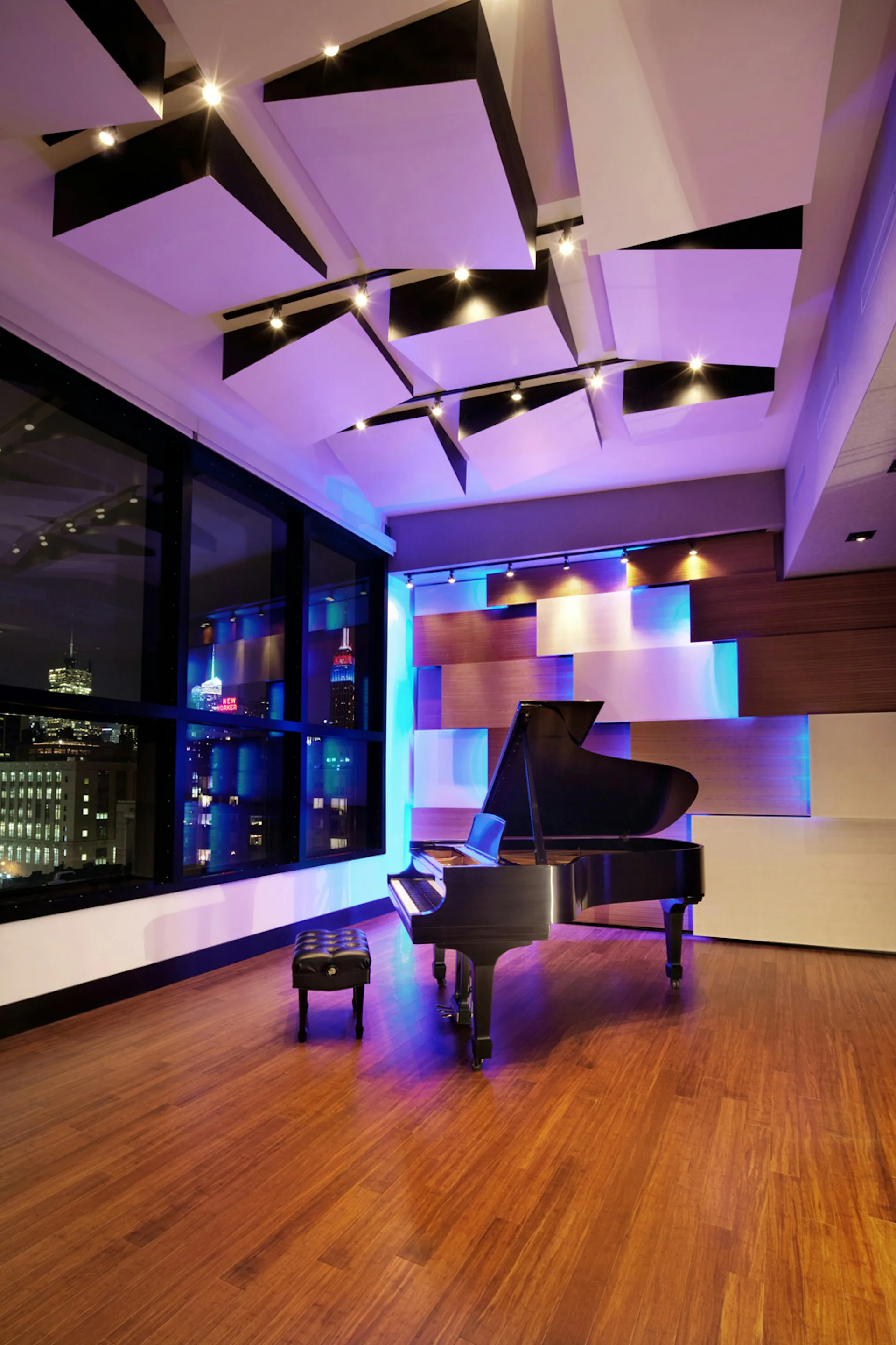 Modern music studio with grand piano, purple and blue ambient lighting, geometric ceiling design with black architectural elements, and city skyline view by Walters-Storyk Design Group.