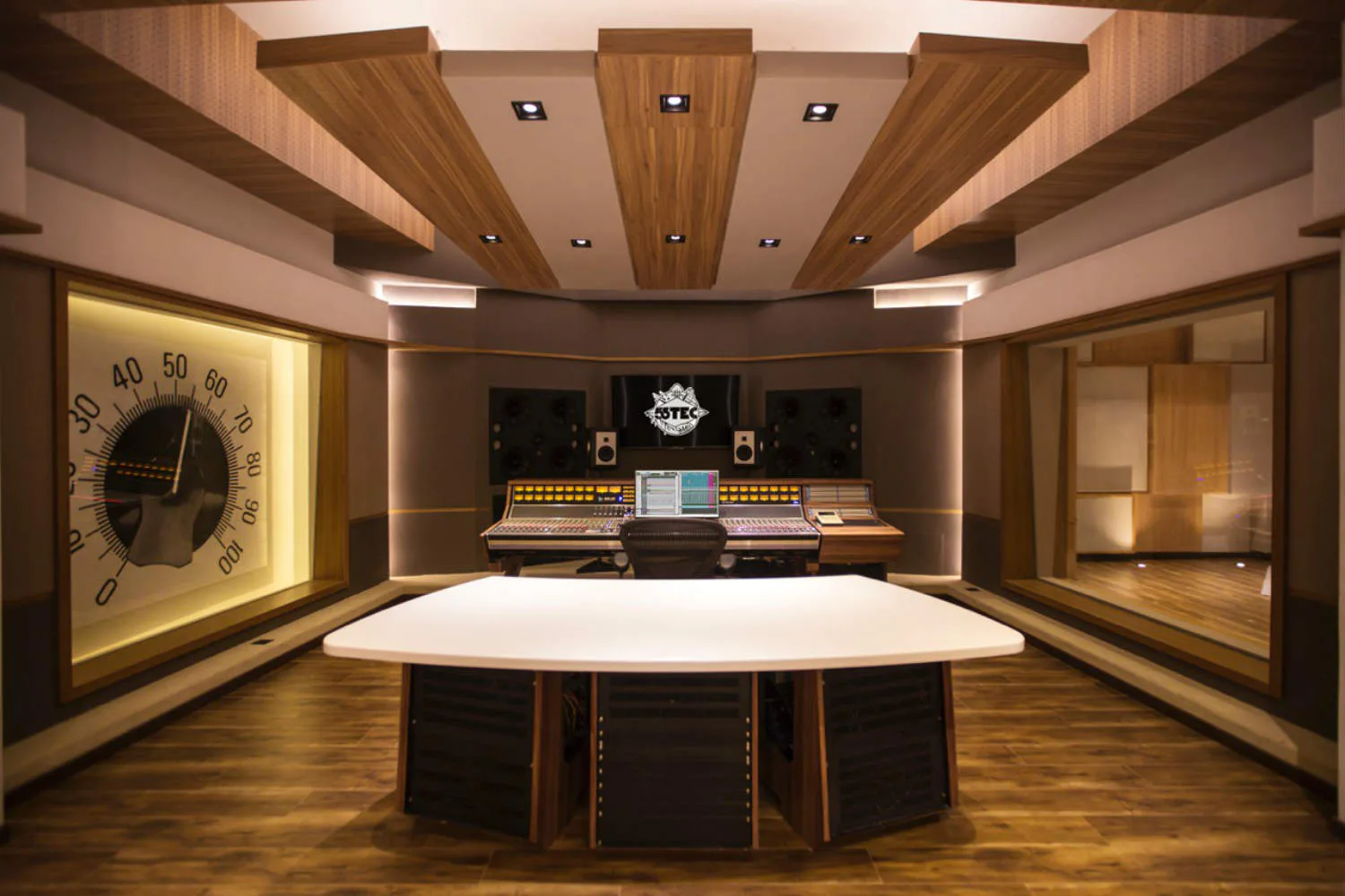 A modern recording studio with a curved white console desk, wooden ceiling details, integrated clock, and symmetrical side rooms with warm wood paneling and professional audio equipment by Walters-Storyk Design Group.