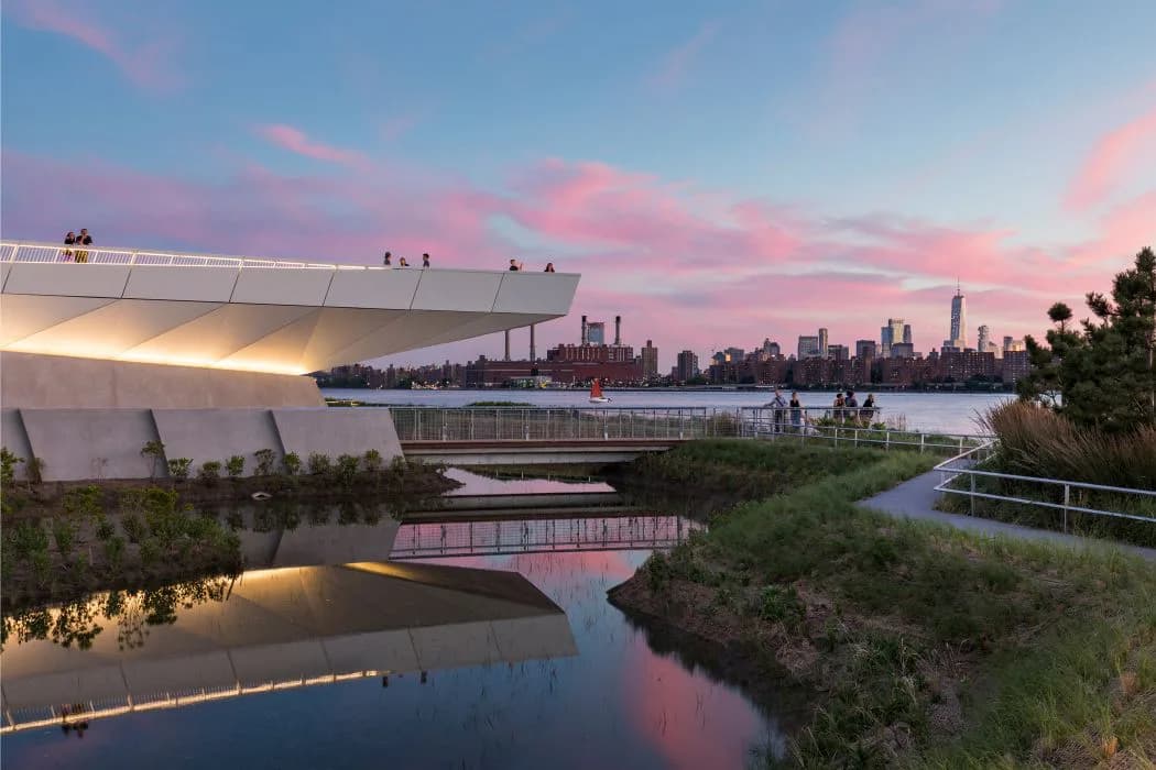 Waterfront park with modern curved pavilion, wetland plantings, and walking paths overlooking Manhattan skyline at dusk by WEISS/MANFREDI.
