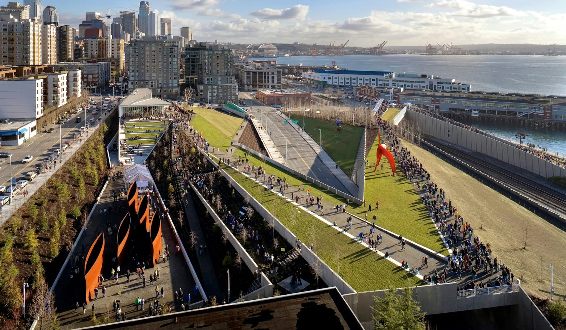 Aerial view of a contemporary waterfront park with tiered green spaces, orange sculptural elements, and integrated pathways overlooking a bay and urban skyline by WEISS/MANFREDI Architecture/Landscape/Urbanism.