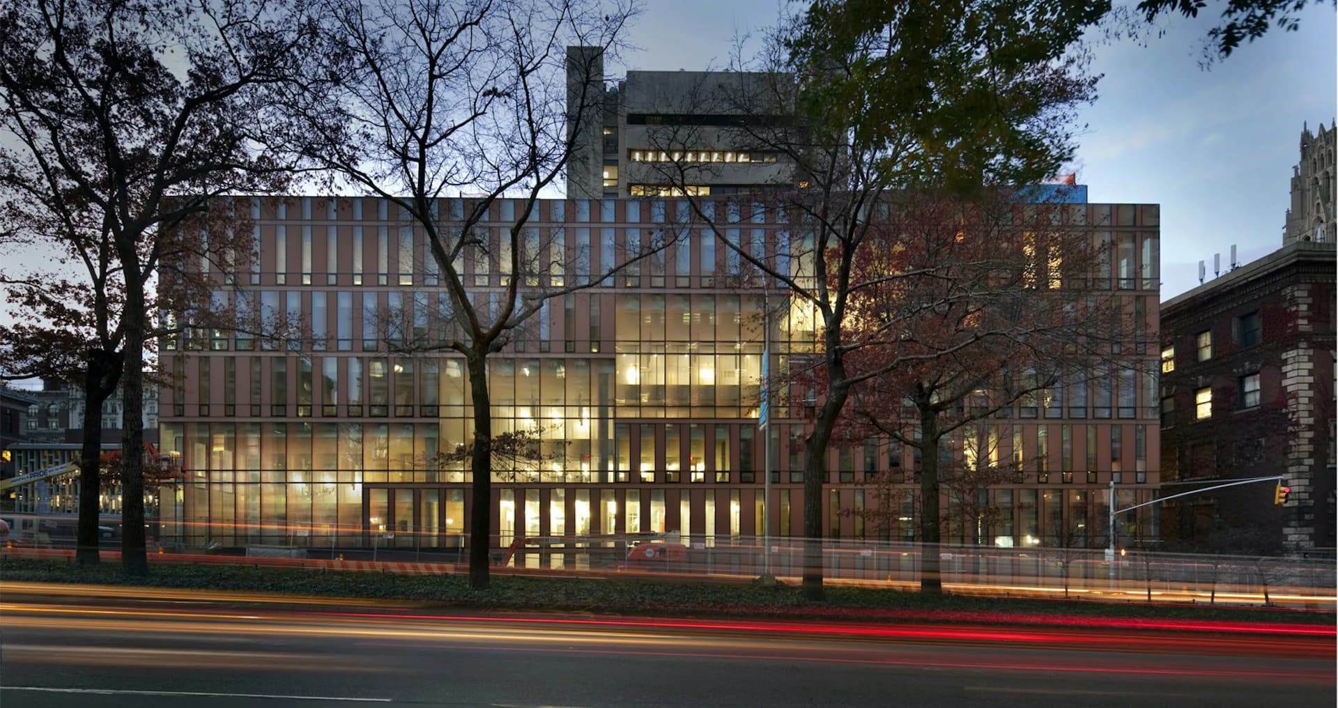 Modern glass and brick building facade illuminated at dusk with surrounding bare trees and historic architecture, designed by WEISS/MANFREDI Architecture/Landscape/Urbanism.