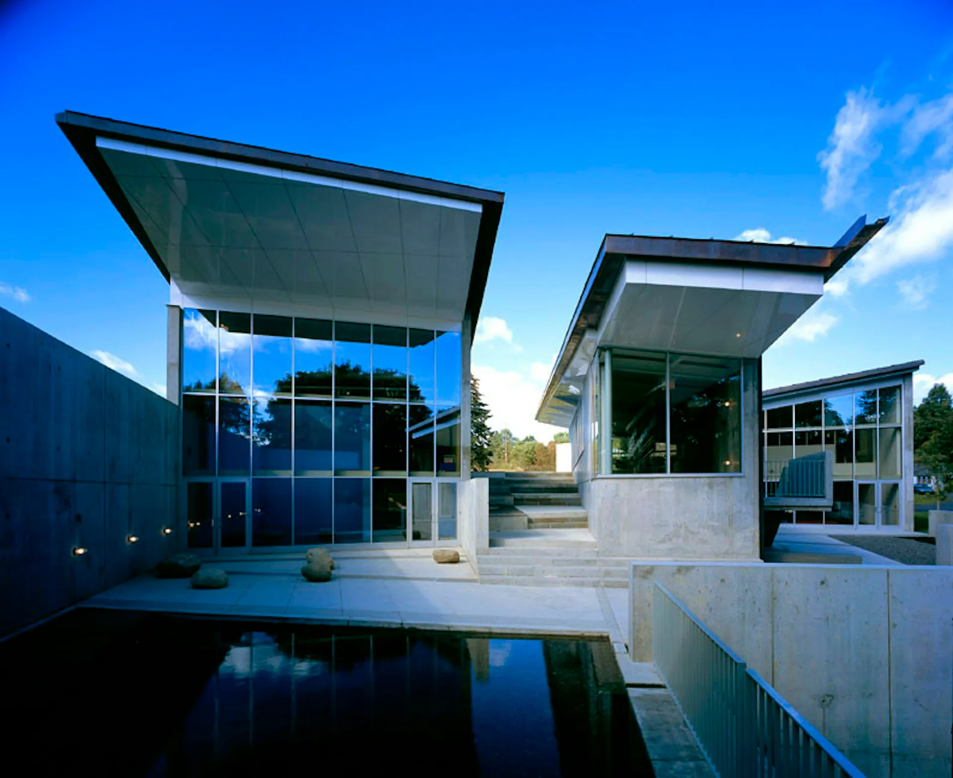 Modern architectural complex with geometric forms, reflective pool, concrete and glass materials, and open courtyard spaces under blue sky by WEISS/MANFREDI Architecture/Landscape/Urbanism.