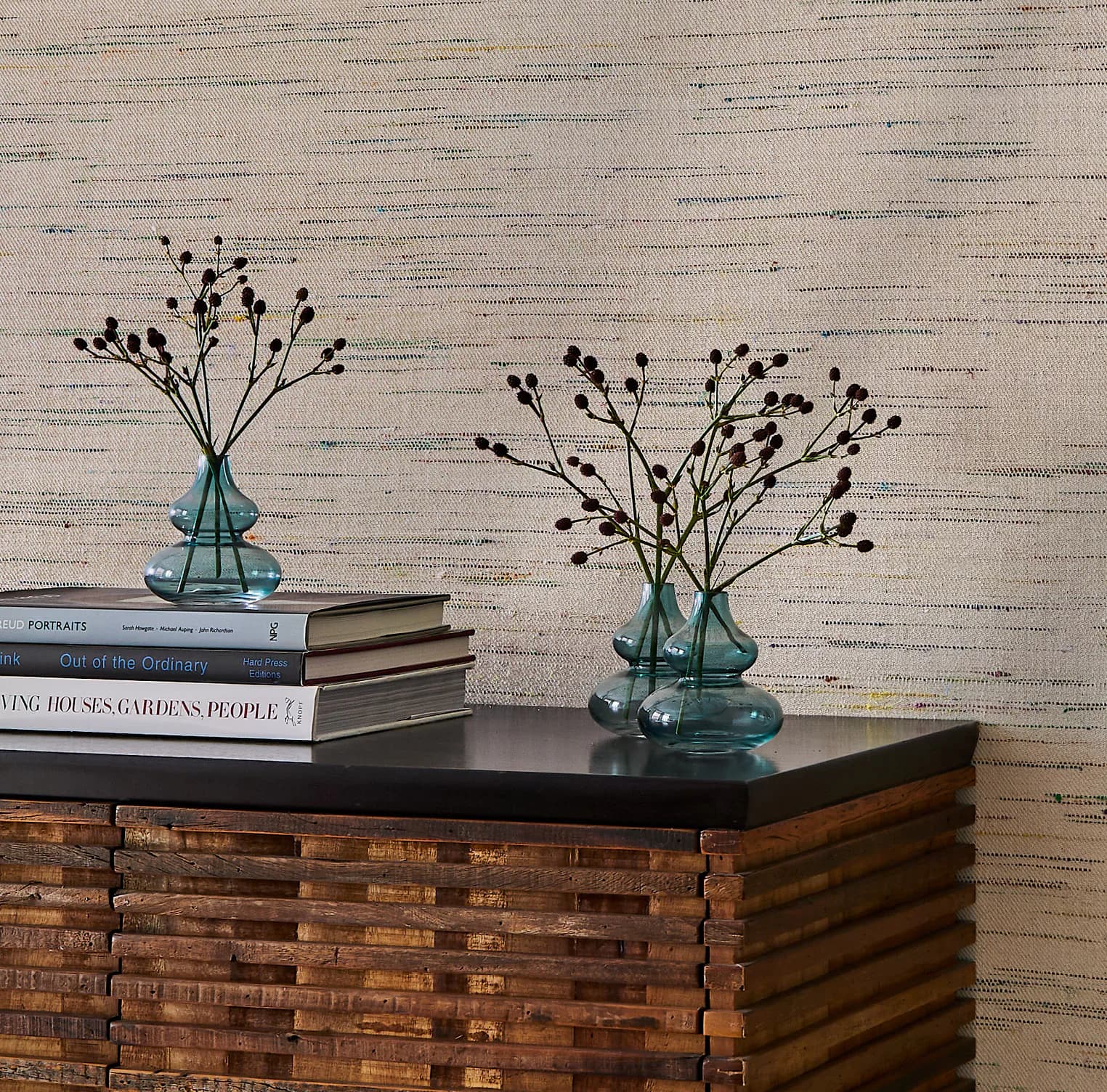 Styled interior vignette featuring a wooden brick-pattern cabinet topped with stacked books, teal glass vessels with dried botanicals, and textured wallpaper backdrop by Weitzner Limited.