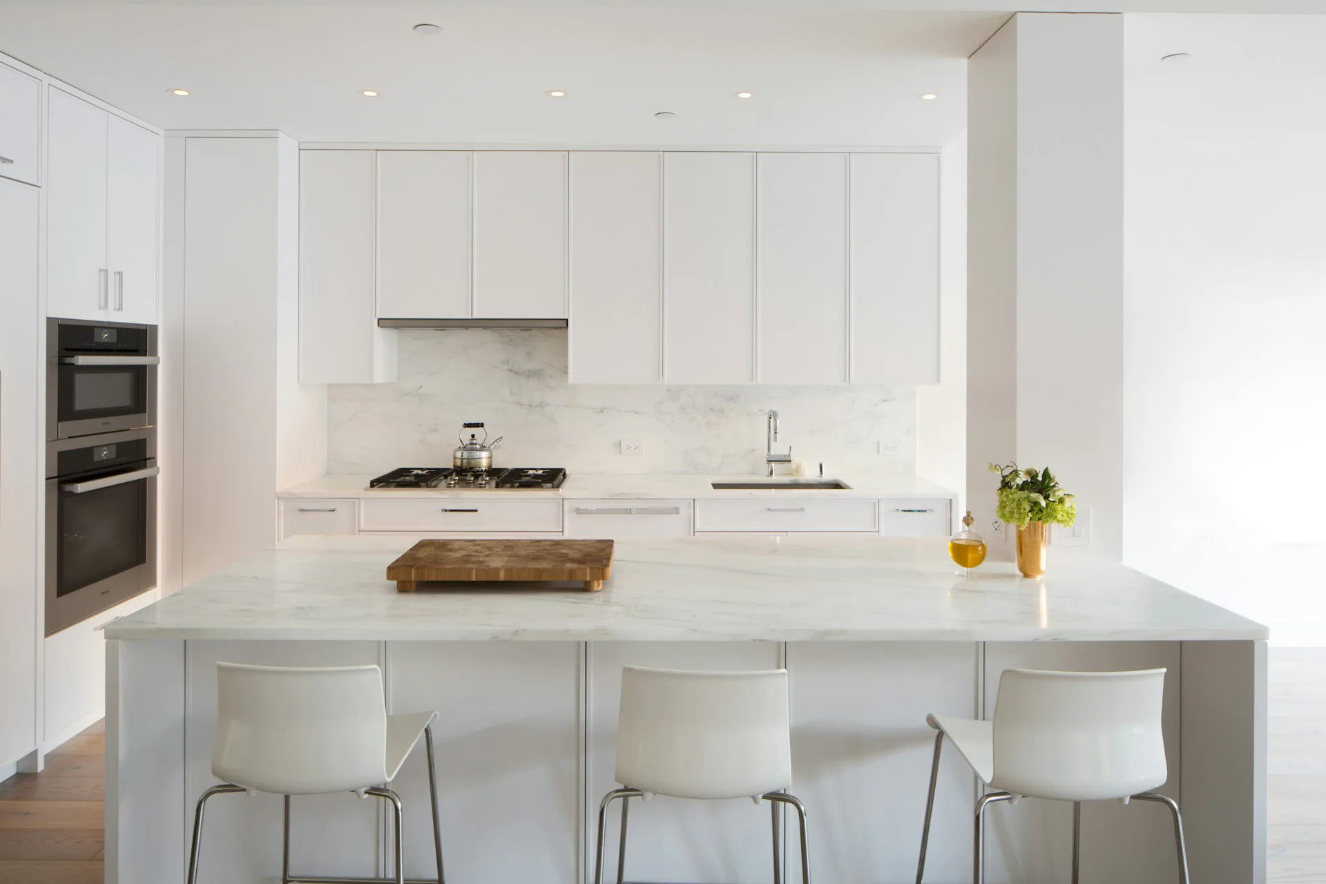 Minimalist white kitchen with waterfall countertop island, integrated appliances, and chrome chairs, designed by Wettling Architects.