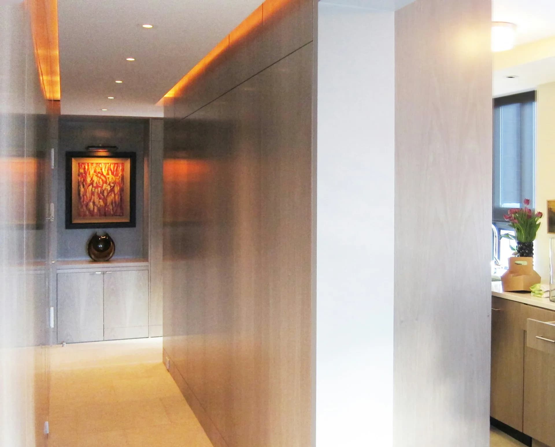 Modern hallway with warm amber ceiling lighting, white walls, polished concrete flooring, and a framed artwork in an illuminated alcove by Wettling Architects.