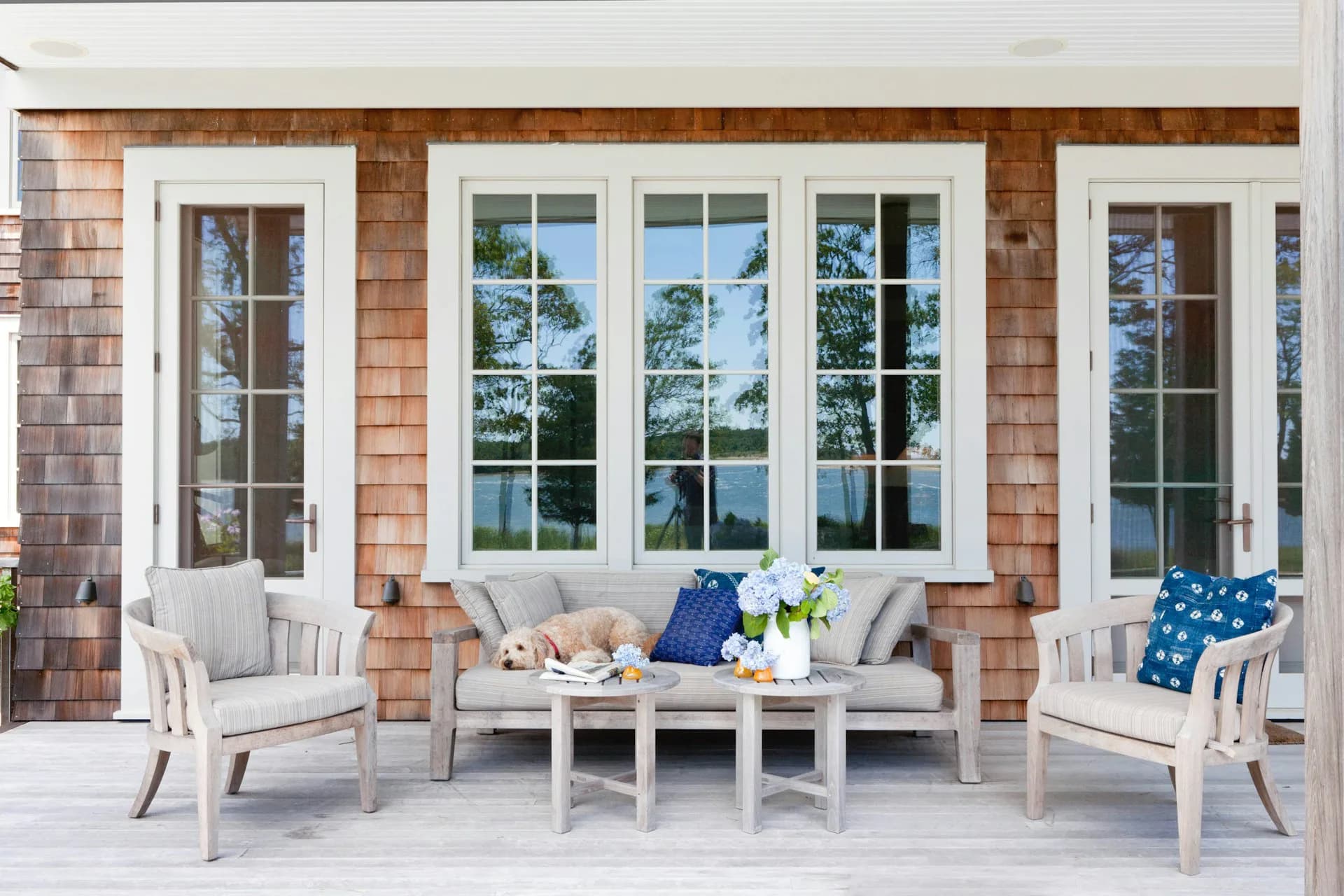 Waterfront porch with brick exterior, white trim windows, gray upholstered seating, and blue accents overlooking water by Wettling Architects.