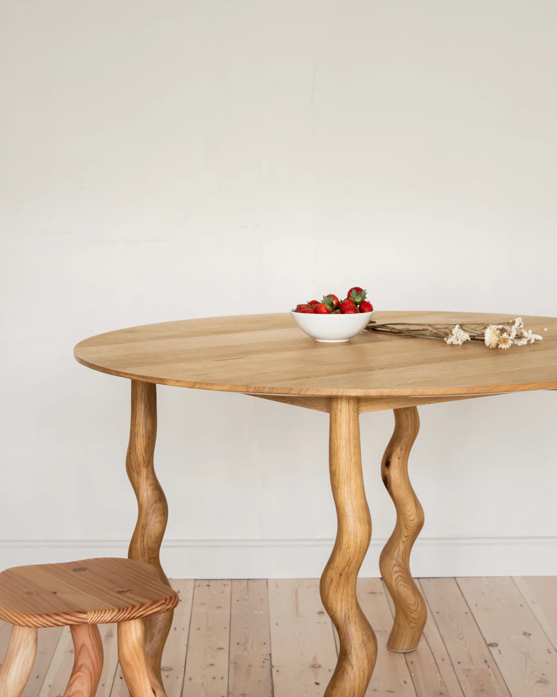 Wooden oval dining table with wavy curved legs and a small matching stool, styled with fresh strawberries and flowers by Wilkinson & Rivera.