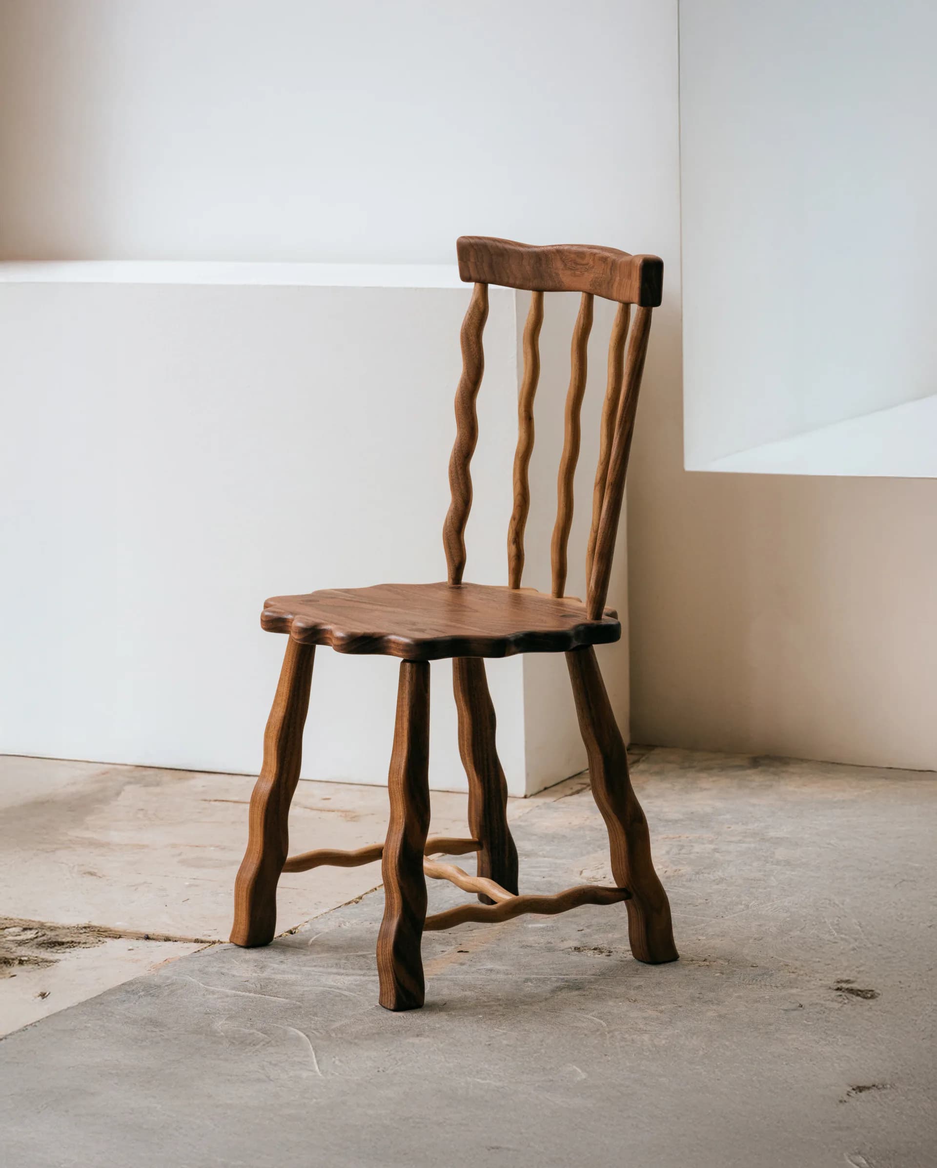 Rustic wooden chair with spindle back and organic curved legs on concrete floor against white wall by Wilkinson & Rivera.
