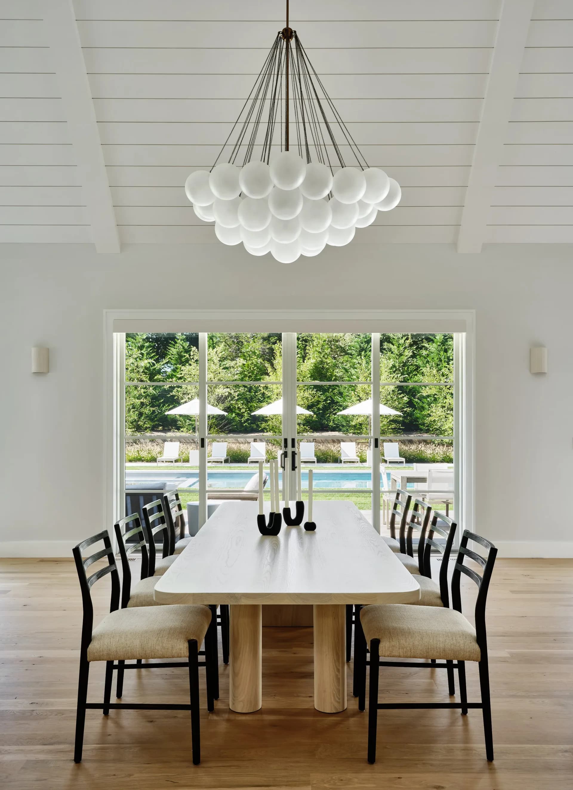 Modern dining room with white shiplap ceiling, sculptural pendant lighting, glass doors overlooking a pool, and black ladder-back chairs by Withers Studio.