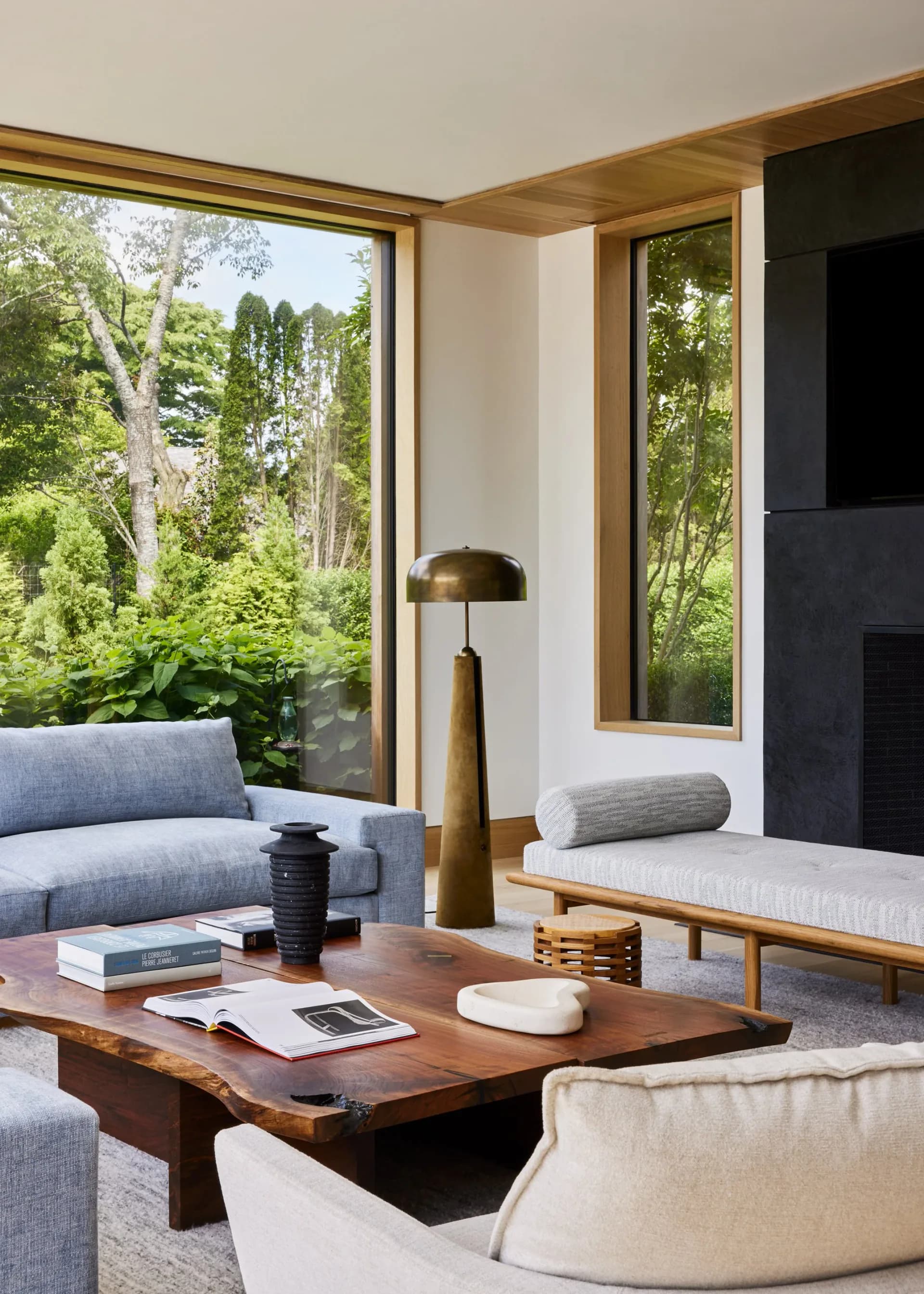 Modern living room with natural wood frames, large windows overlooking trees, light gray upholstered furniture, wooden coffee table, and brass pendant lamp by Withers Studio.