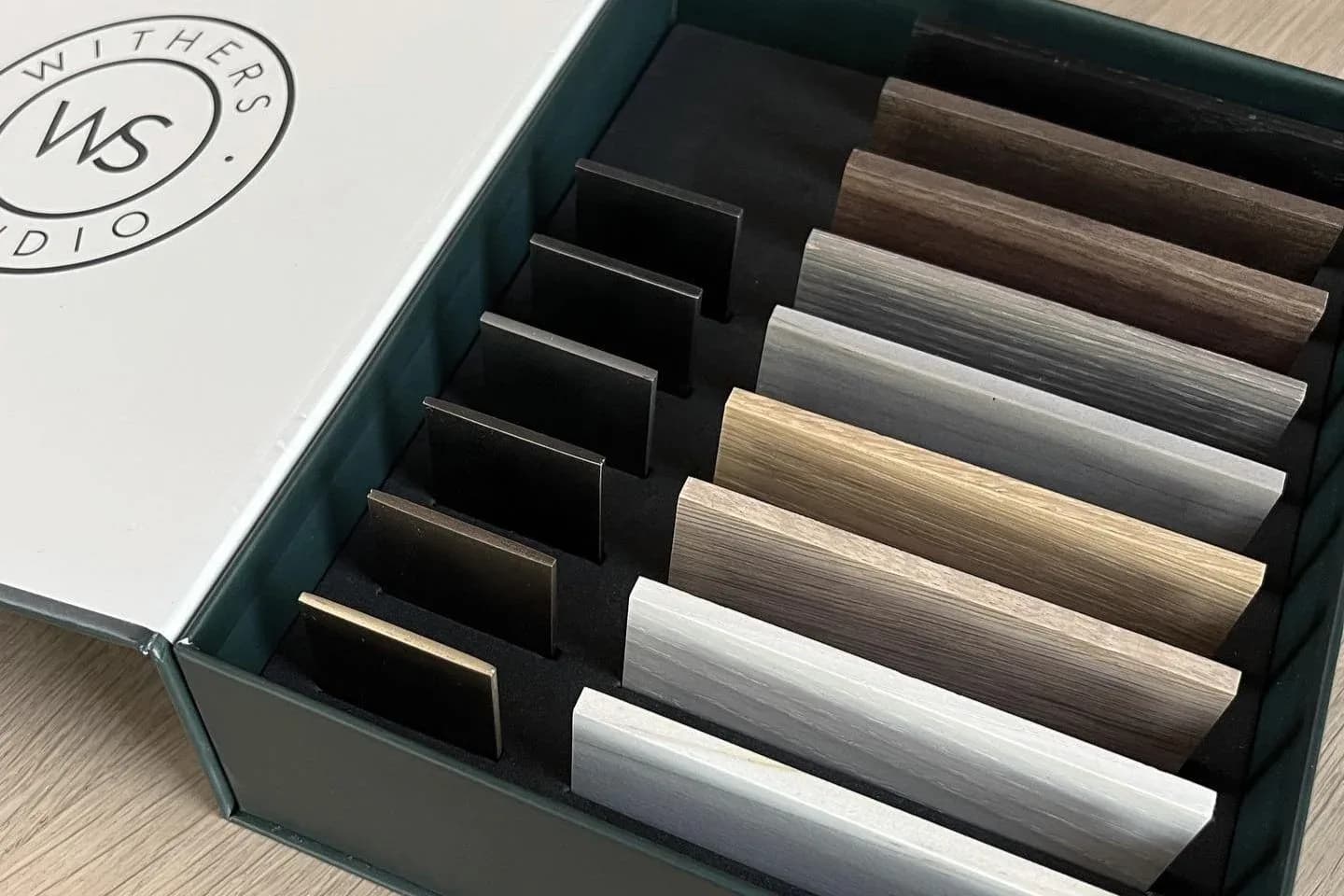 Open sample box displaying material swatches in organized rows of black, wood, and neutral tones by Withers Studio.