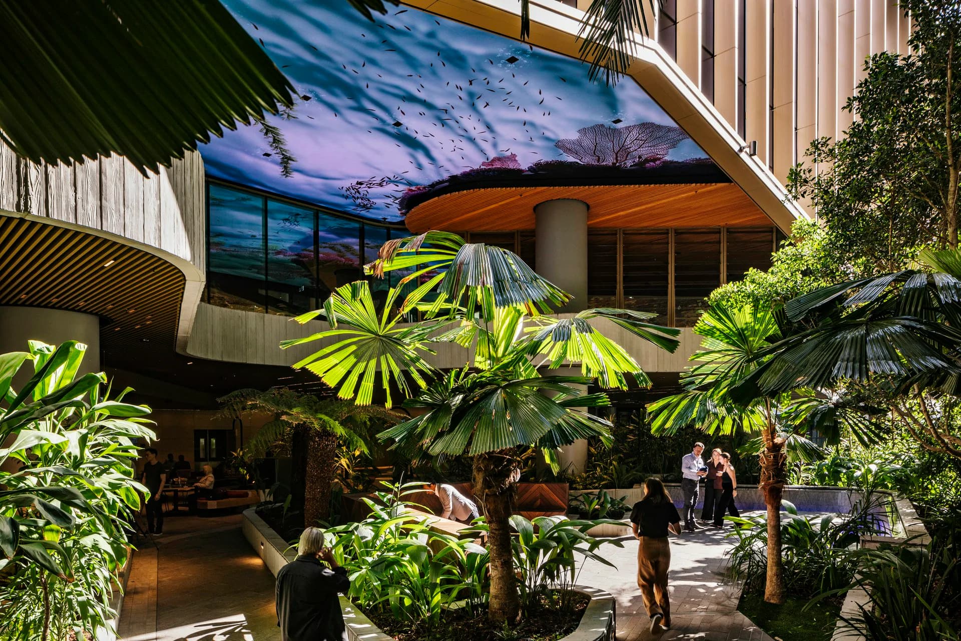 Modern multi-level atrium with dramatic sky-lit ceiling, lush tropical plantings, and contemporary architecture featuring warm wood and metal elements by Woods Bagot.