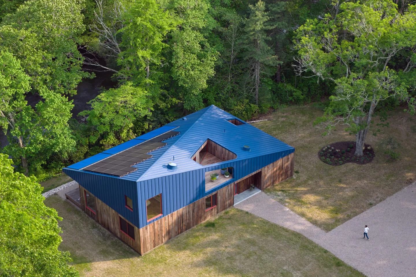 Aerial view of a modernist pavilion with blue metal roof and weathered wood siding surrounded by forest and lawn, by WORKac.