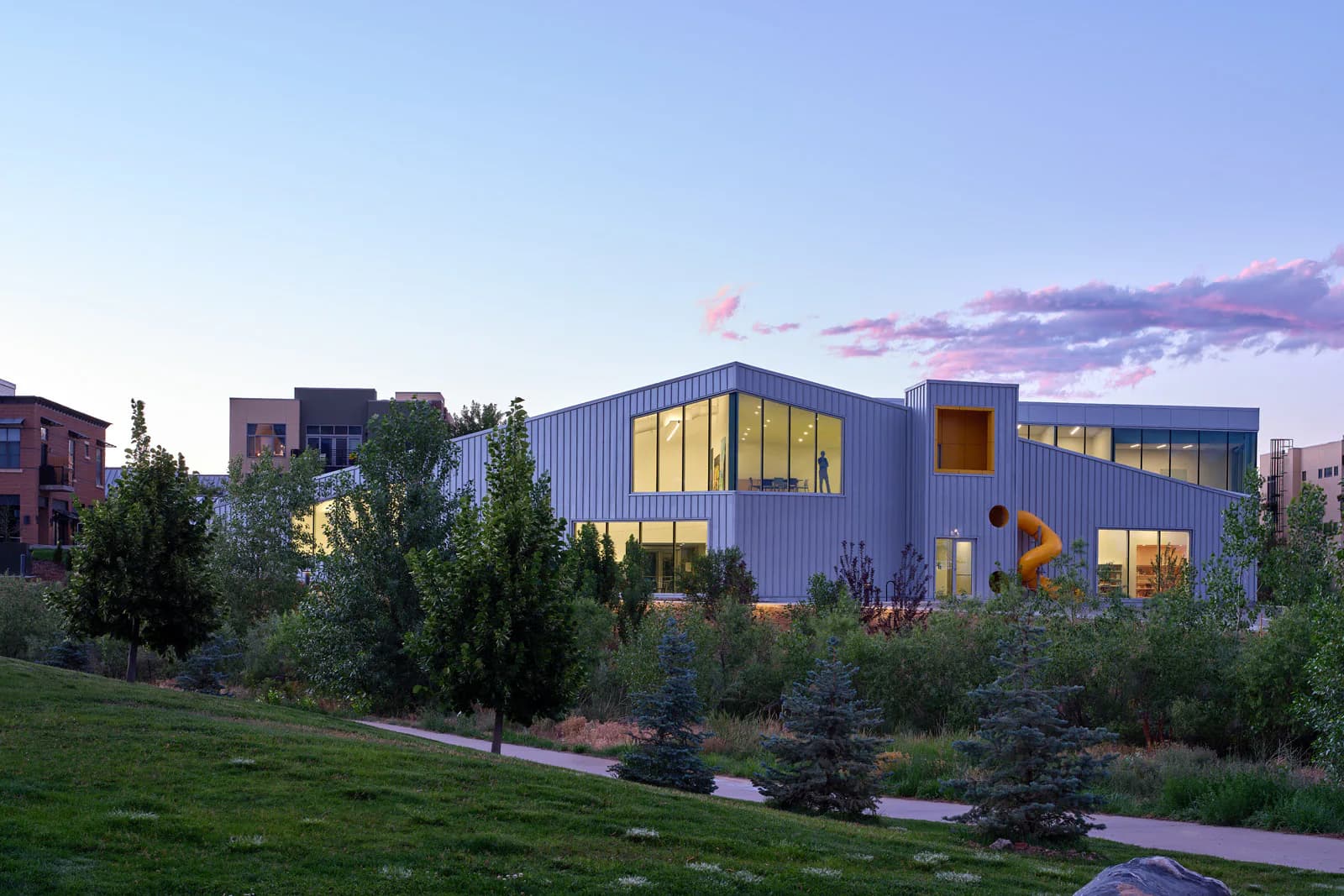 Contemporary corrugated metal building with yellow-lit windows set among landscaped grounds and trees at dusk, designed by WORKac.