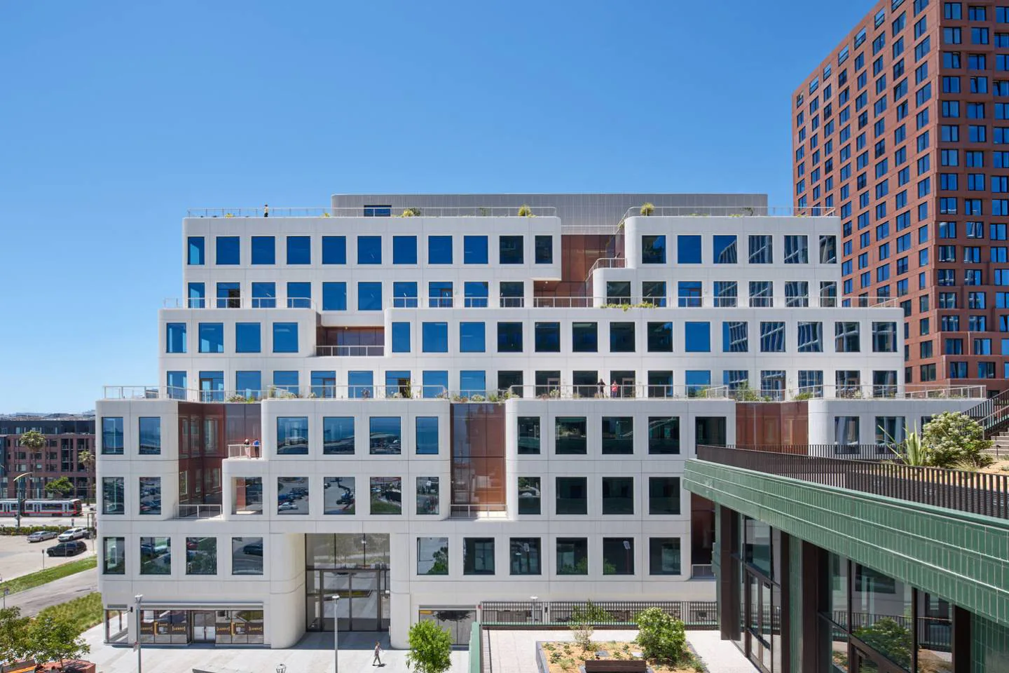 Contemporary multi-story mixed-use building with white and brick façade, regular window grid, ground-floor retail, and green pedestrian bridge by WORKac.