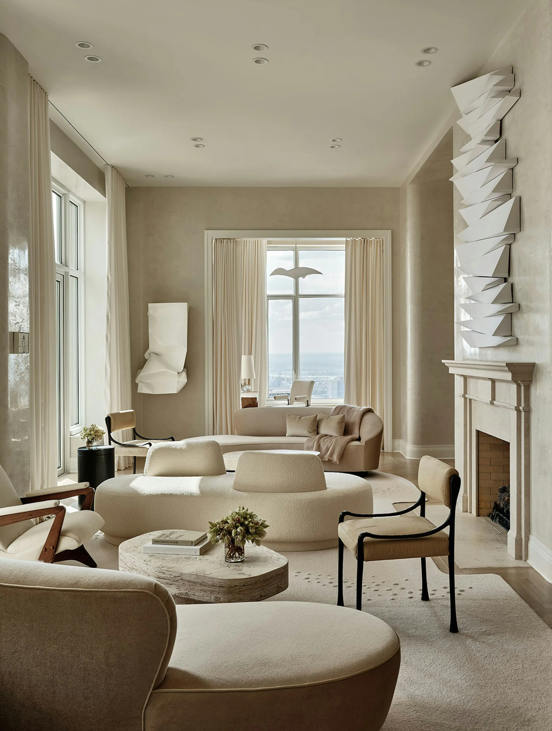Luxurious living room with curved cream furniture, travertine coffee table, fireplace, and floor-to-ceiling windows overlooking water by Workshop/APD.