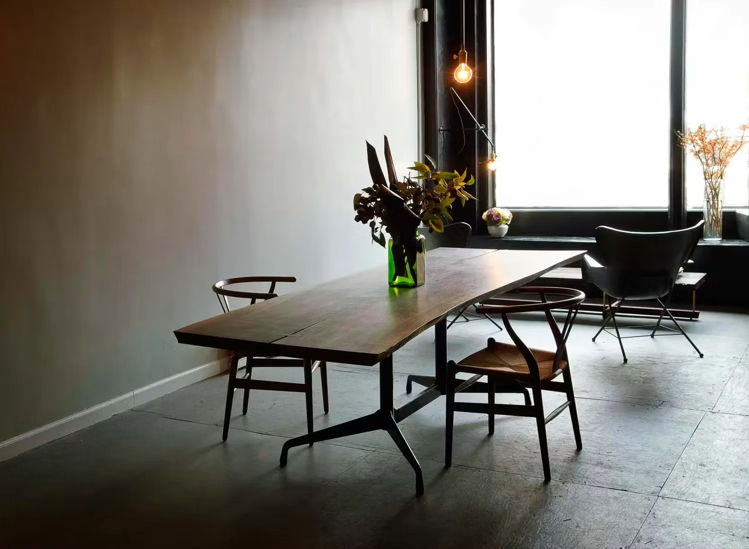 Modern dining space with dark wood table, mid-century chairs, concrete flooring, and large windows with natural light, by Workstead.