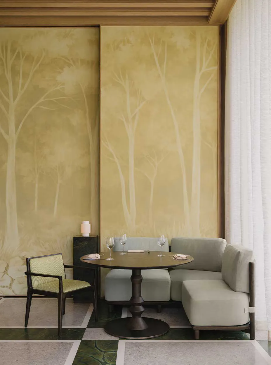 Intimate dining nook featuring a round pedestal table, cream banquette seating, and a single accent chair, set against golden tree-patterned wall panels by Yabu Pushelberg.