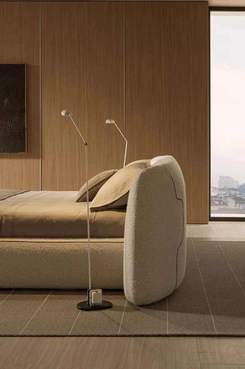 Minimalist bedroom with warm wood-paneled walls, cream upholstered armchair, brass floor lamps, and city views by Yabu Pushelberg.