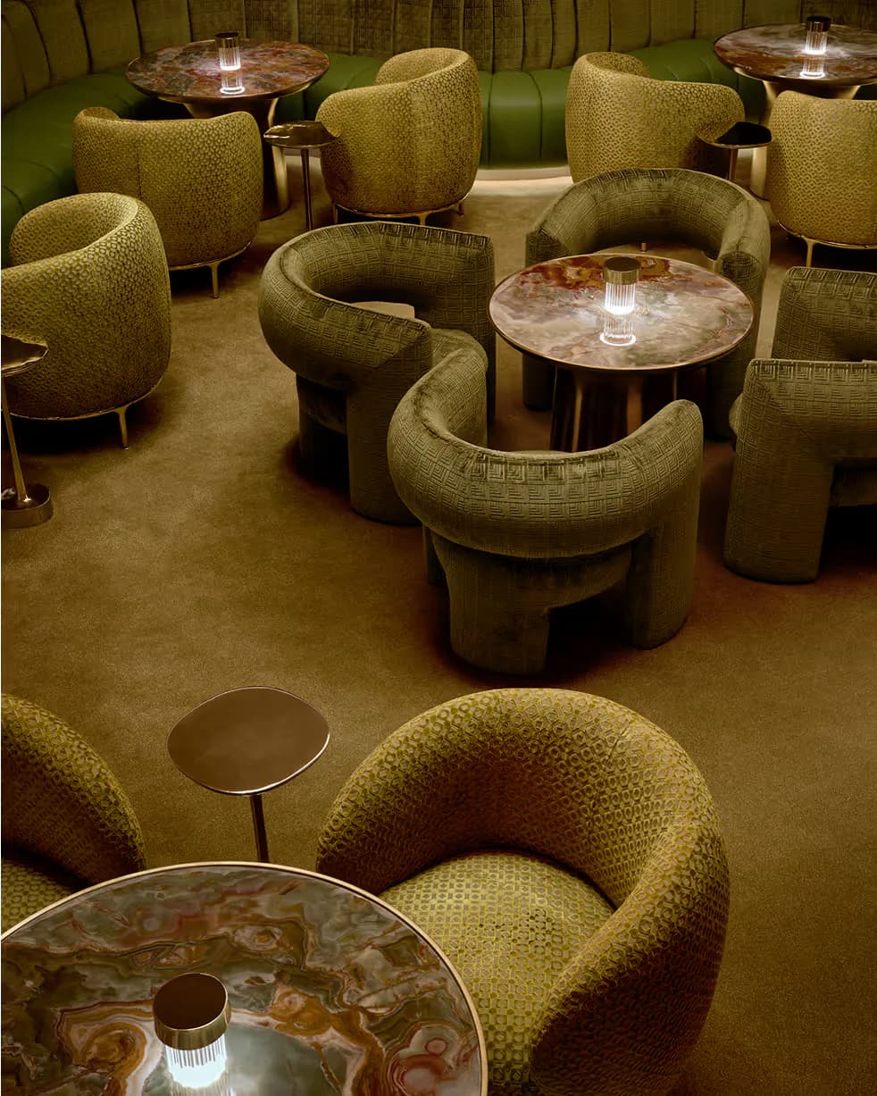 Lounge area with curved olive-green upholstered seating, polished stone side tables with integrated lighting, and warm tan flooring, designed by Yabu Pushelberg.