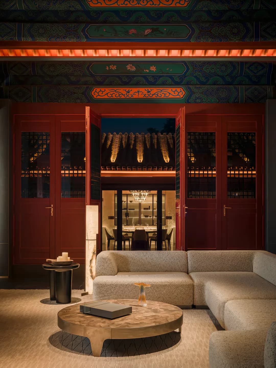 A luxurious lounge features a curved beige sectional sofa, round wooden coffee table, and cylindrical side table facing ornate red doors with traditional Asian architectural details and decorative panels above by Yabu Pushelberg.