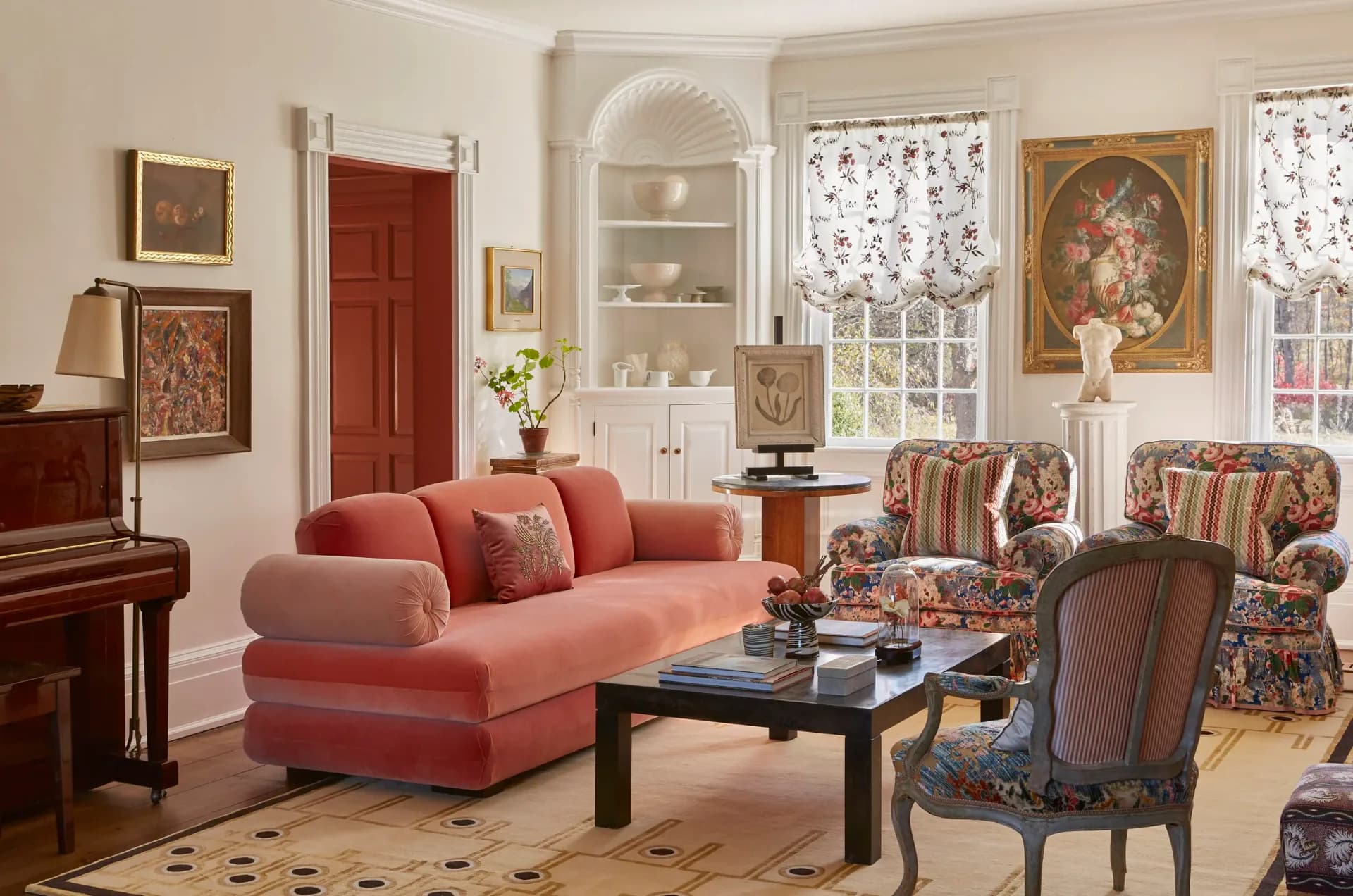 A traditional living room features a coral-pink sectional sofa, patterned accent chairs, black coffee table, and built-in cabinetry with shelving, designed by Young Huh Interior Design.