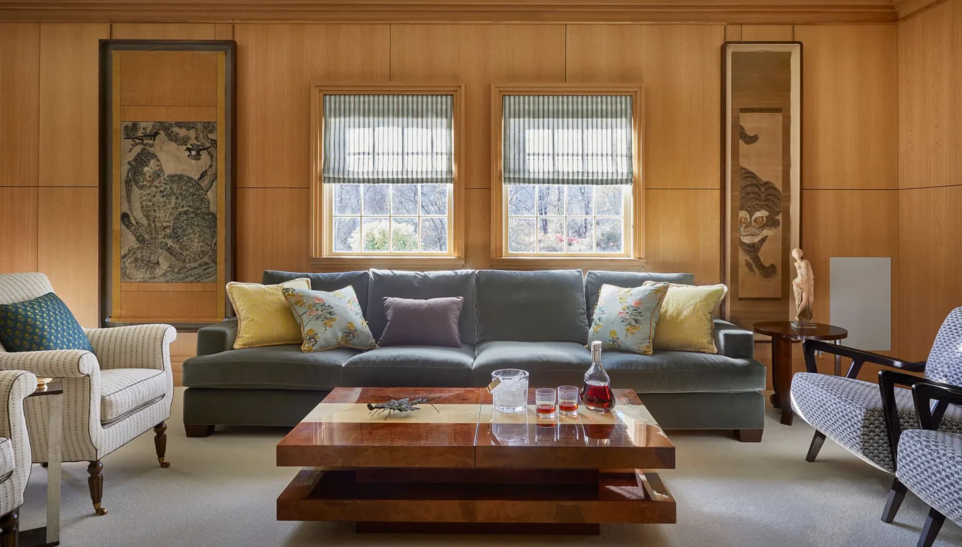 Warm wood-paneled living room with gray sectional sofa, colorful accent pillows, wooden coffee table, and upholstered accent chairs by Young Huh Interior Design.