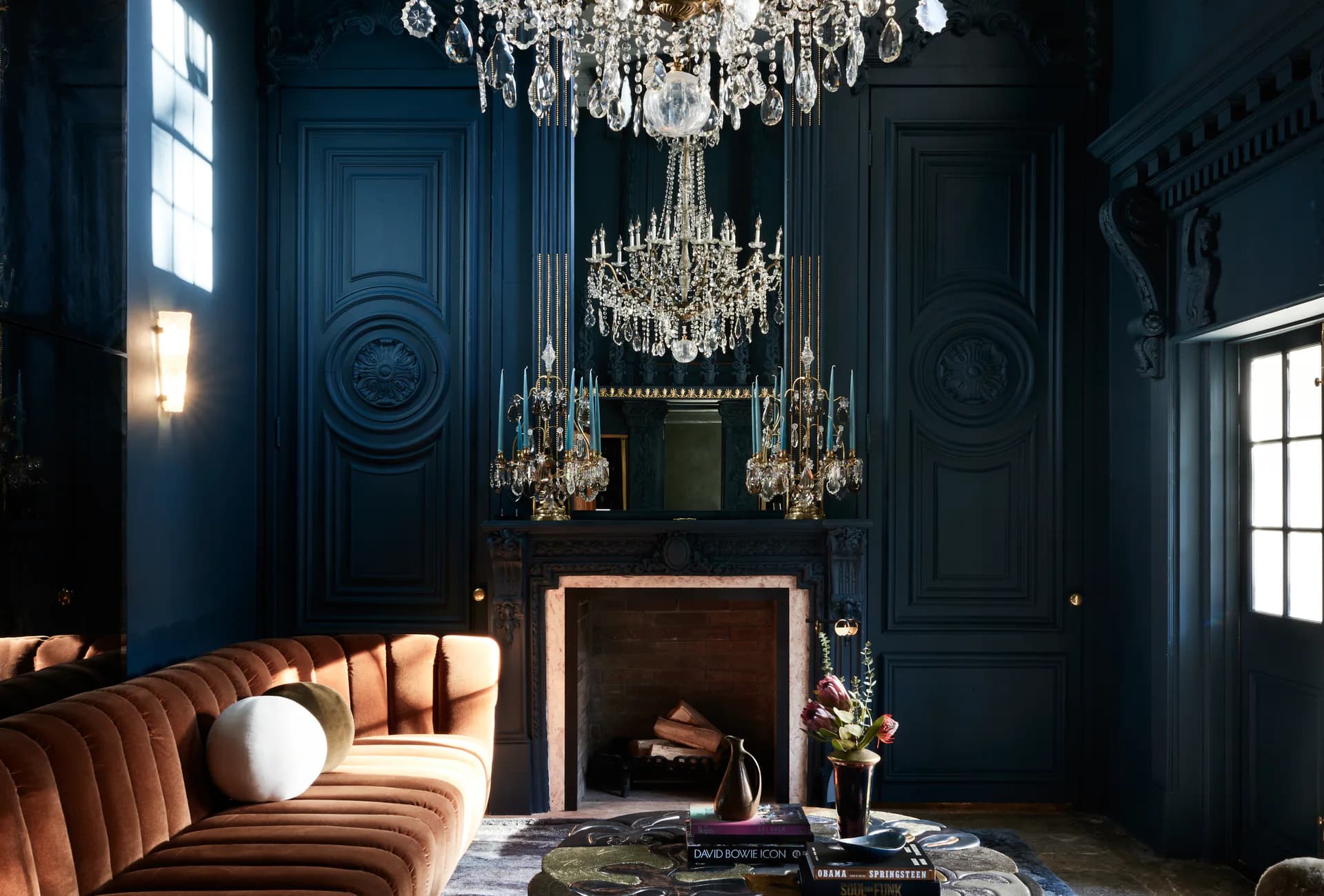 Luxurious blue-walled room with curved cognac leather sofa, crystal chandeliers, fireplace with brass surround, and classical architectural details by Young Projects.