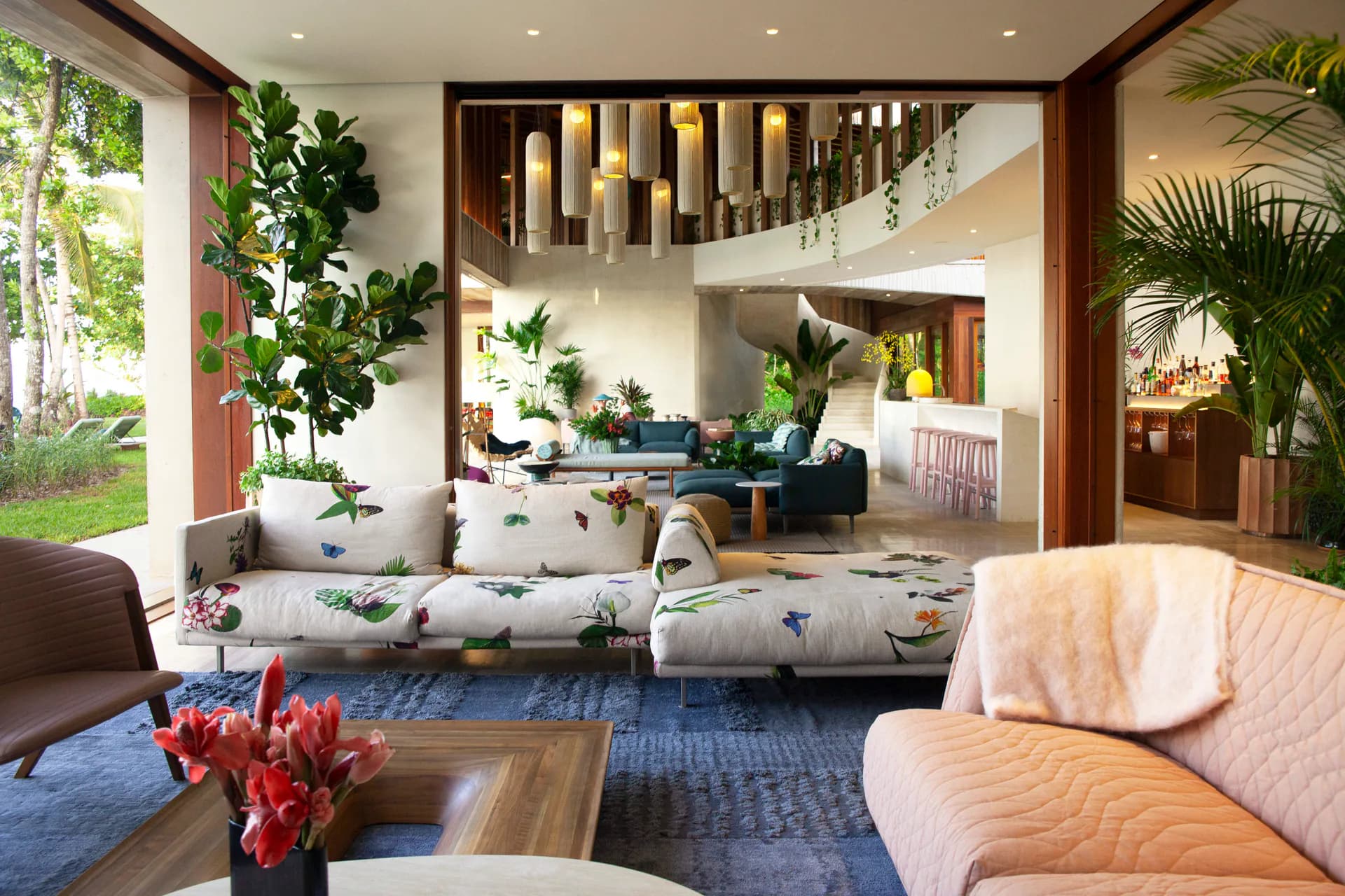 Multi-level residential interior with natural wood framing, blue area rug, cream upholstered seating, and abundant greenery throughout by Young Projects.