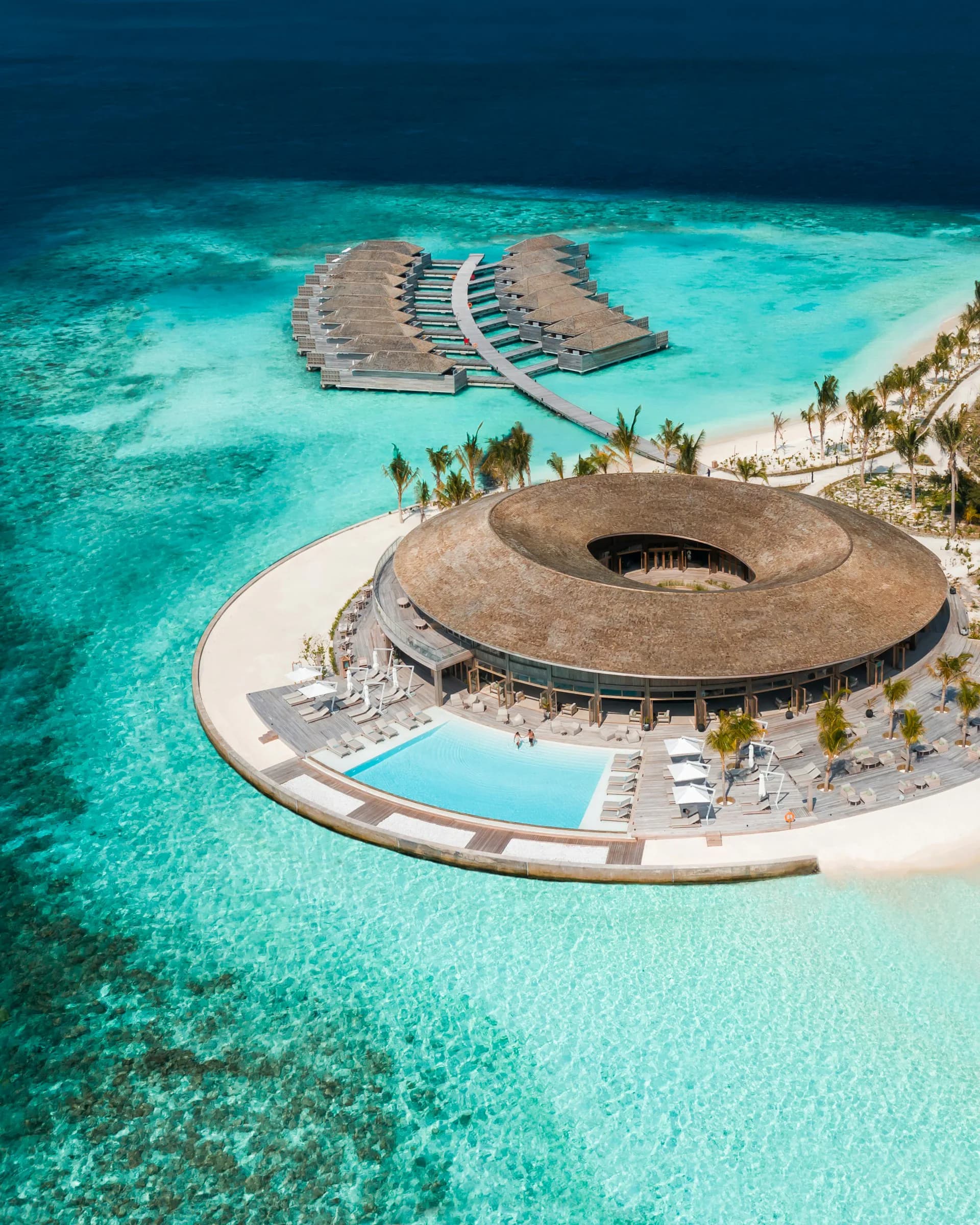 Aerial view of a circular resort building with a distinctive torus design featuring a central atrium, surrounded by curved beach loungers and a lagoon pool, set on turquoise waters with palm trees by Yuji Yamazaki Architecture.