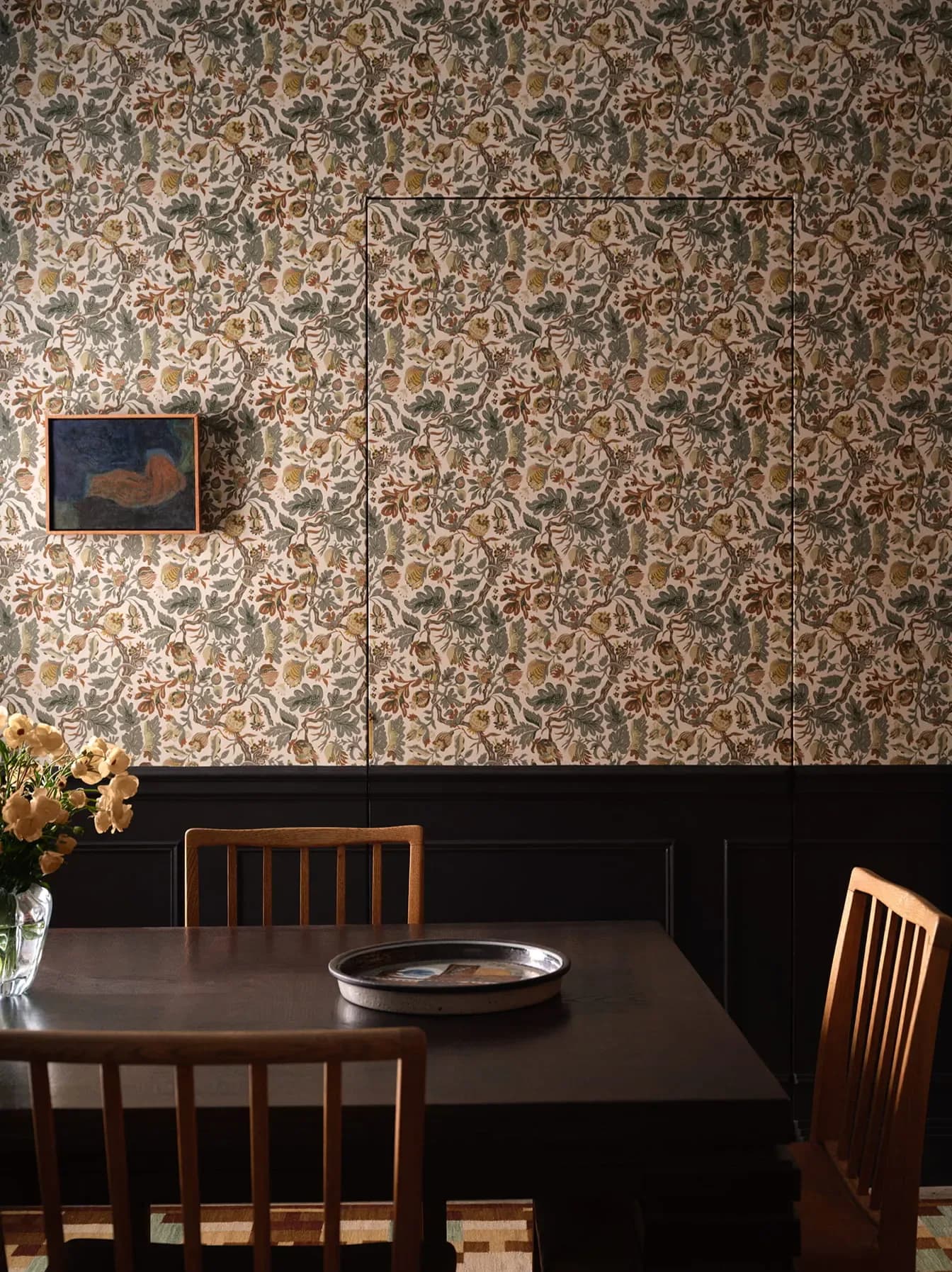 Dining room with ornate damask wallpaper, black wainscoting, dark wood table and spindle-back chairs, small framed artwork by Zak + Fox.