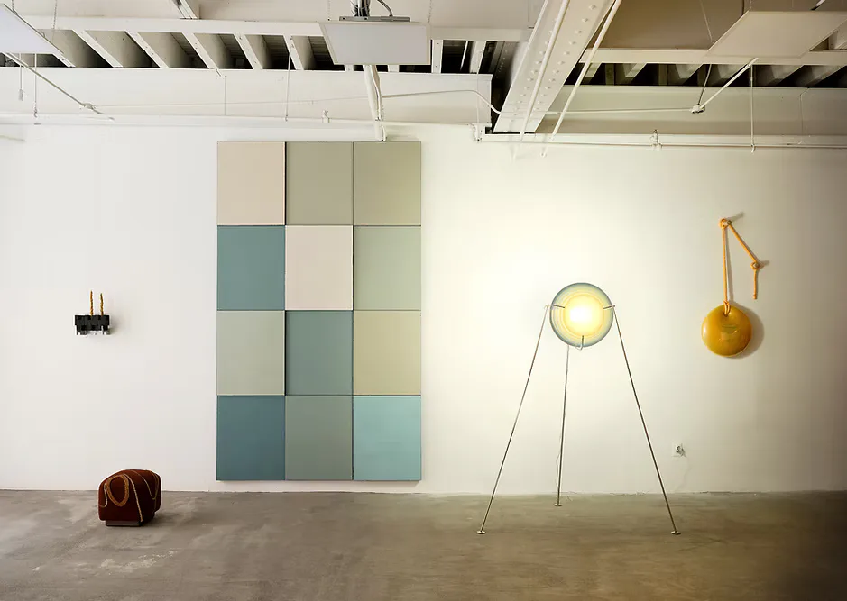 Minimalist gallery installation featuring a teal and cream modular wall panel, pendant lighting fixtures, and sculptural elements on concrete flooring by ZAROLAT.